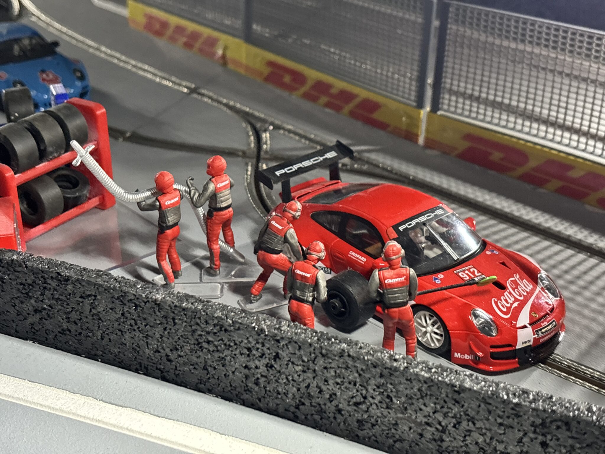 Gros plan sur un arrêt aux stands miniature du circuit Good Time Racing, avec une Porsche rouge et des mécaniciens figurines en combinaison rouge changeant les pneus